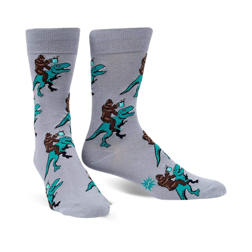 Pair of grey crew socks featuring a repeating pattern of Bigfoot riding a teal T-Rex and holding a mug. 