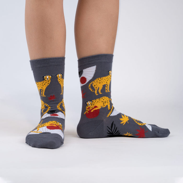 A person is modeling a pair of gray socks with a pattern of red Japanese maple leaves, yellow and white fans, and black and yellow spotted leopards holding red balls in their mouths.