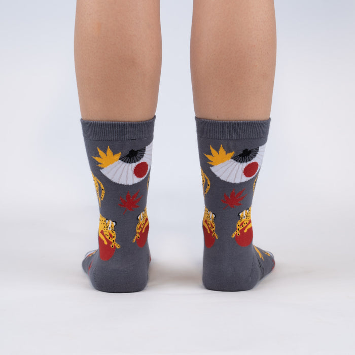 A person is modeling a pair of gray socks with a pattern of red Japanese maple leaves, yellow and white fans, and black and yellow spotted leopards holding red balls in their mouths.