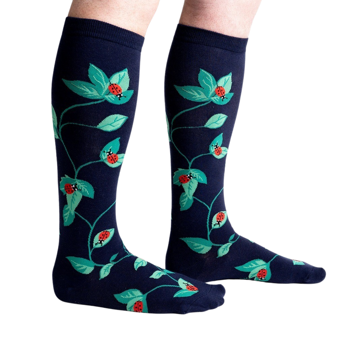 A pair of dark blue knee-high socks with a repeating pattern of red ladybugs and green leaves.