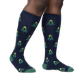 Lifestyle shot showing a person's legs wearing the Bigfoot tree socks. The socks are navy blue with a repeating pattern of green Bigfoot figures and lime green Christmas trees.