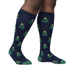 Lifestyle shot showing a person's legs wearing the Bigfoot tree socks. The socks are navy blue with a repeating pattern of green Bigfoot figures and lime green Christmas trees.
