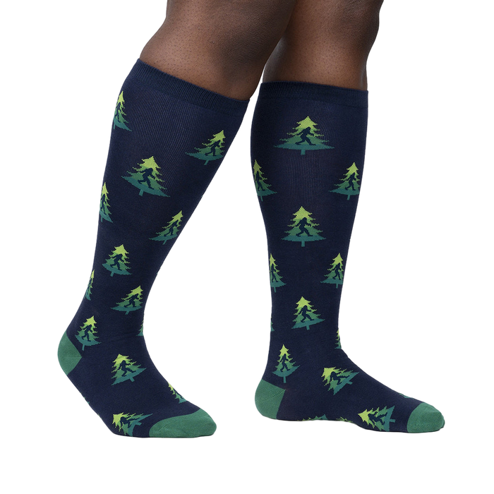 Lifestyle shot showing a person's legs wearing the Bigfoot tree socks. The socks are navy blue with a repeating pattern of green Bigfoot figures and lime green Christmas trees.