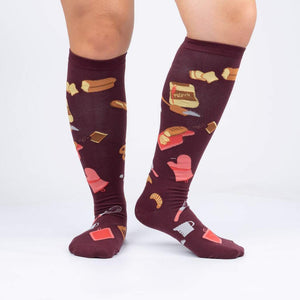 A pair of knee-high socks with a baking theme. The socks are maroon with various baking-related items on them, such as bread, pastries, and kitchen utensils.