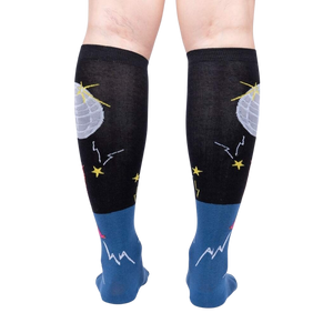 A pair of black knee-high socks with a blue section at the ankle and a pink heel and toe. There is a pattern of stars and moons on the black section and a mountain pattern on the blue section.