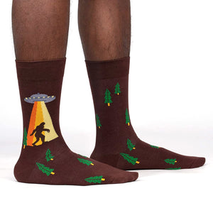 A pair of brown socks with a pattern of green pine trees. On the left sock is a picture of Bigfoot walking with a UFO beaming up above him.