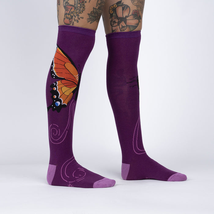 A pair of purple knee-high socks with a butterfly pattern on the back. The butterfly has orange and black wings with white markings. The socks are being modeled by a person with tattoos on their legs.
