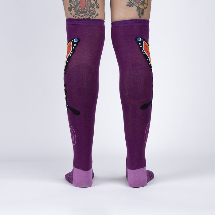 A pair of purple knee-high socks with a butterfly pattern on the back. The butterfly has orange and black wings with white markings. The socks are being modeled by a person with tattoos on their legs.
