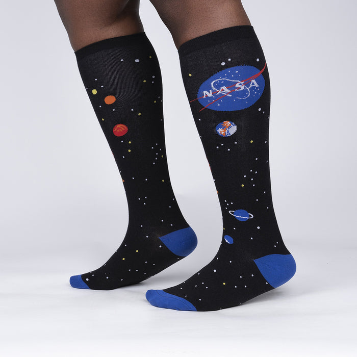 A pair of black knee-high socks with a NASA logo on the left leg and a pattern of stars on both legs. The socks are being worn by a person who is standing with their back to the camera.