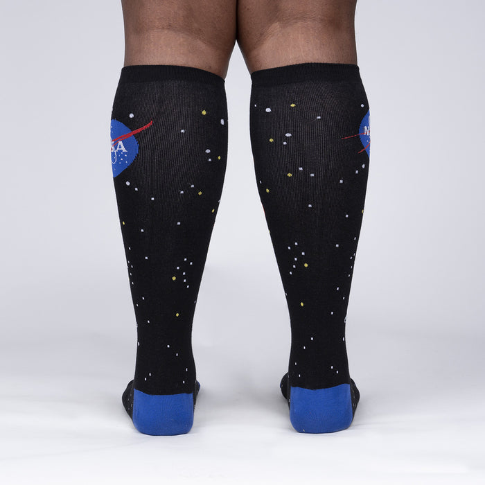 A pair of black knee-high socks with a NASA logo on the left leg and a pattern of stars on both legs. The socks are being worn by a person who is standing with their back to the camera.