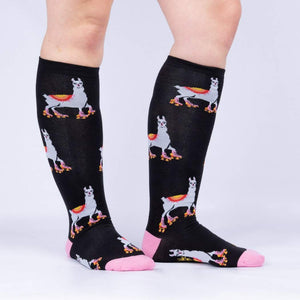 A pair of black knee-high socks with a pattern of cartoon llamas wearing roller-skates in pink, yellow, blue and orange. The socks are shown on the back of a person's legs from the knees down.