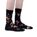 Lifestyle shot showcasing the 'Let That Shiitake Go' socks on a model's feet.  The socks are black with a mushroom and pun design.