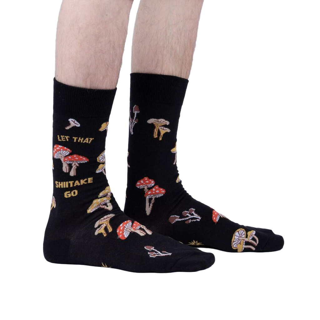 Lifestyle shot showcasing the 'Let That Shiitake Go' socks on a model's feet.  The socks are black with a mushroom and pun design.