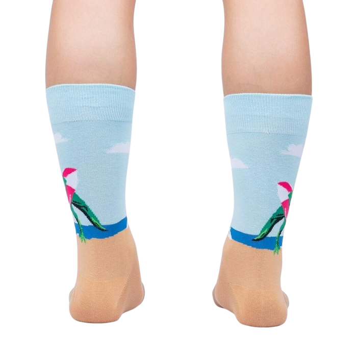 A pair of light blue socks with a pattern of pink flamingos wearing Santa hats on a sandy beach with the ocean in the background.