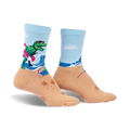 A pair of crew socks showcasing a T-Rex surfing on a beach.  The socks are light blue and tan with green, pink, and white accents.