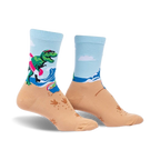 A pair of crew socks showcasing a T-Rex surfing on a beach.  The socks are light blue and tan with green, pink, and white accents.