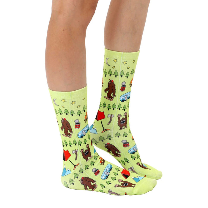 A pair of green socks with a pattern of cartoon Bigfoot creatures in various camping situations.