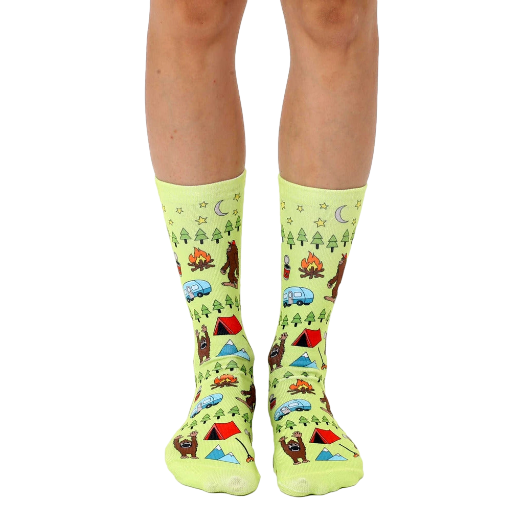Lime green crew socks with a whimsical camping scene featuring Bigfoot, tents, campfires, and other outdoor elements. [lf]