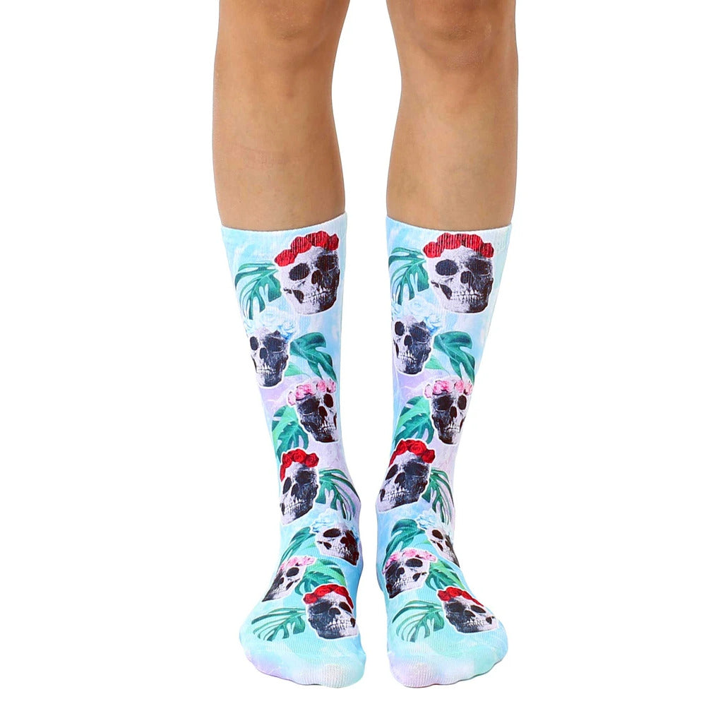 Pair of crew socks in pastel blue, featuring a whimsical pattern of skulls wearing floral crowns and tropical leaves. [lf]