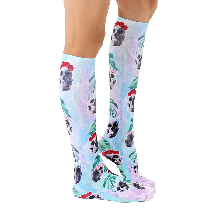 A pair of knee-high socks with a pattern of skulls wearing red flower crowns on a blue and white tie-dye background.