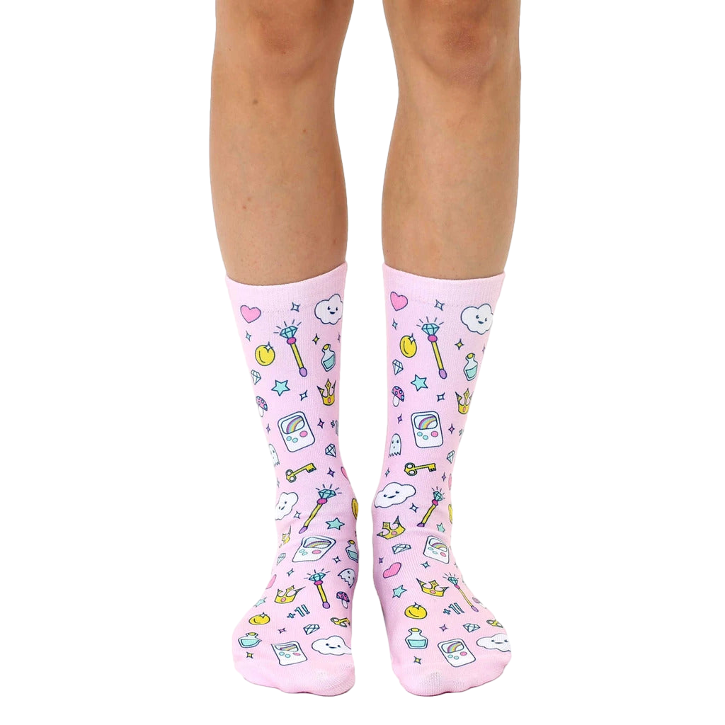 A pair of women's crew socks on a white background.  They're pink with a fun pattern of cartoon magic wands, crowns, and game controllers. [lf]