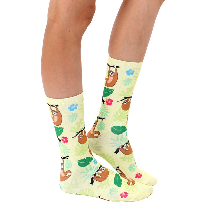 A pair of yellow socks with a pattern of sloths hanging from branches. The sloths are brown with black claws and surrounded by green leaves and pink flowers.