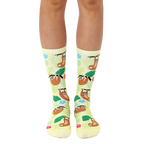 A pair of crew socks on a model's legs. Pale yellow background with cartoon sloths and tropical flowers. [lf]