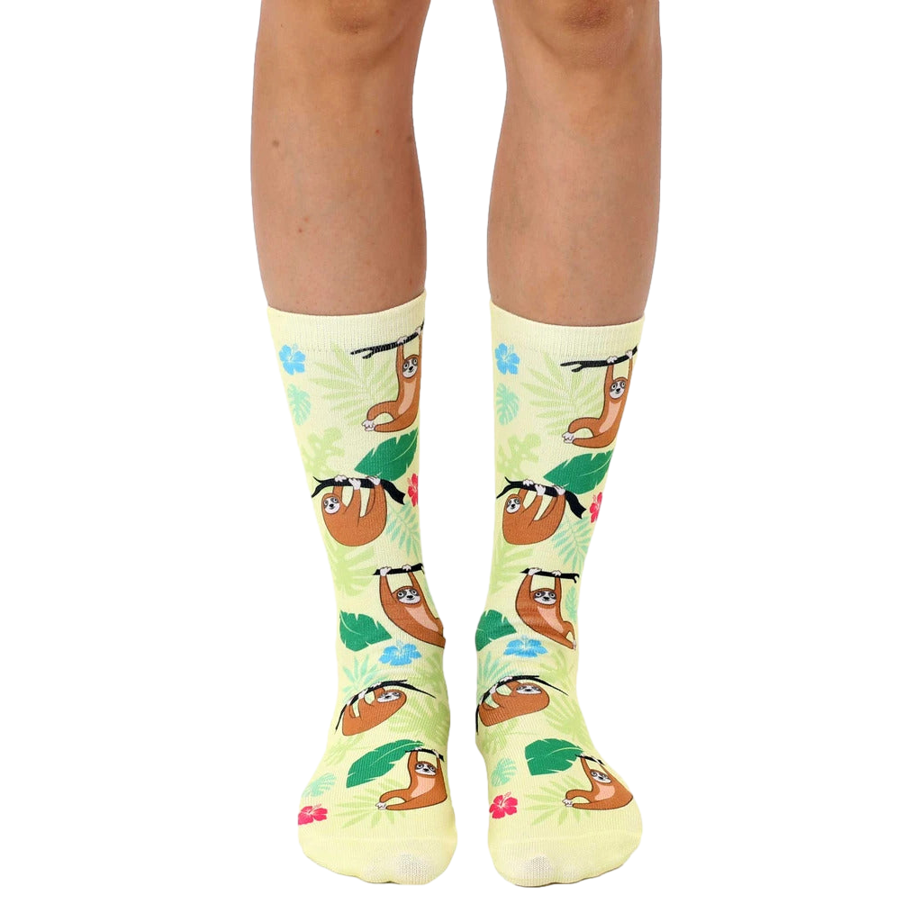 A pair of crew socks on a model's legs. Pale yellow background with cartoon sloths and tropical flowers. [lf]