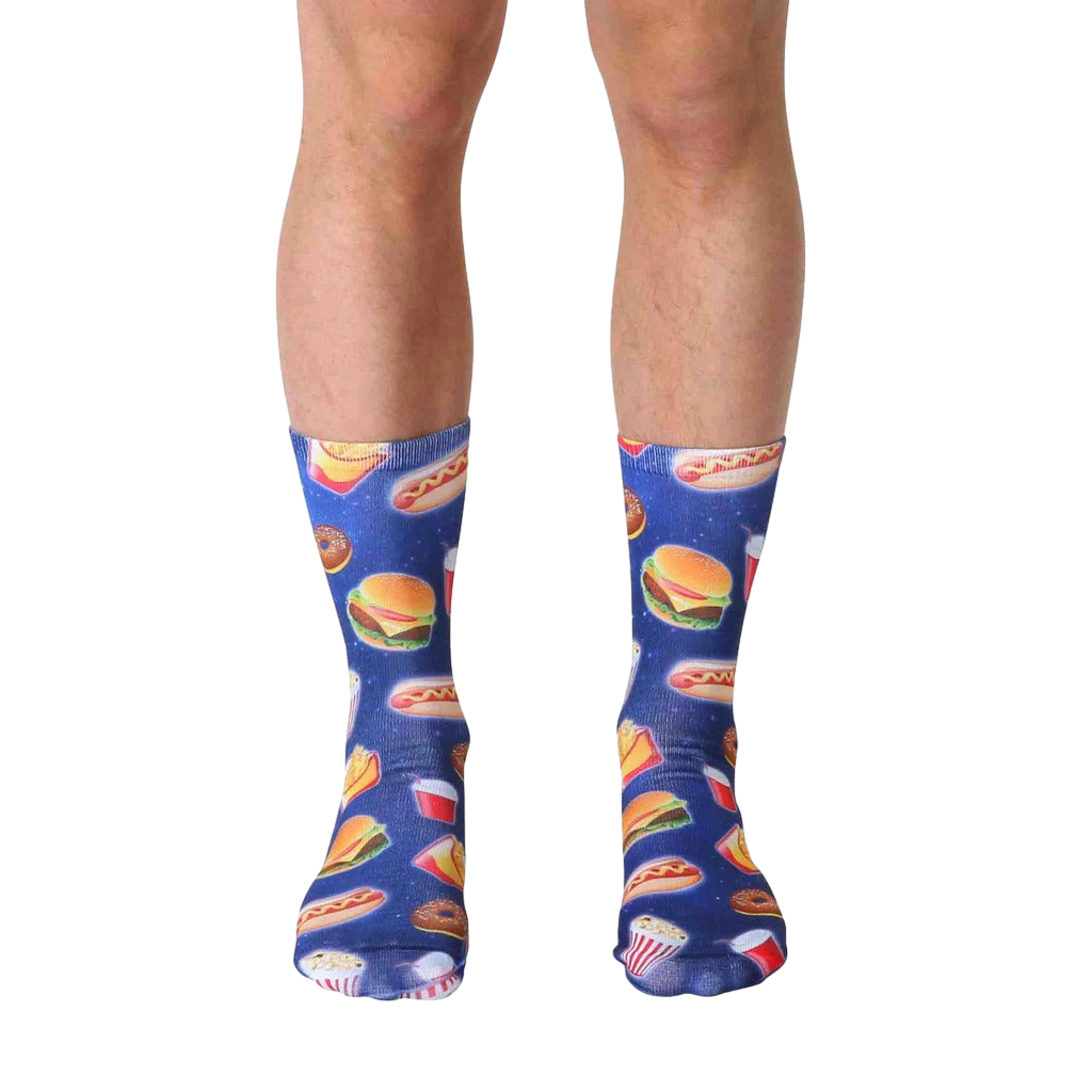 A pair of blue crew socks with a fun pattern of fast food items like burgers, fries, and hot dogs against a galaxy-printed background. [lf]