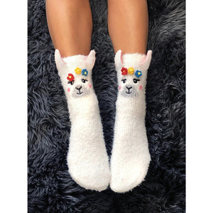 A pair of white fuzzy socks with 3D llama faces on them. The llamas have pink cheeks and are wearing headbands with yellow, pink, and blue flowers.