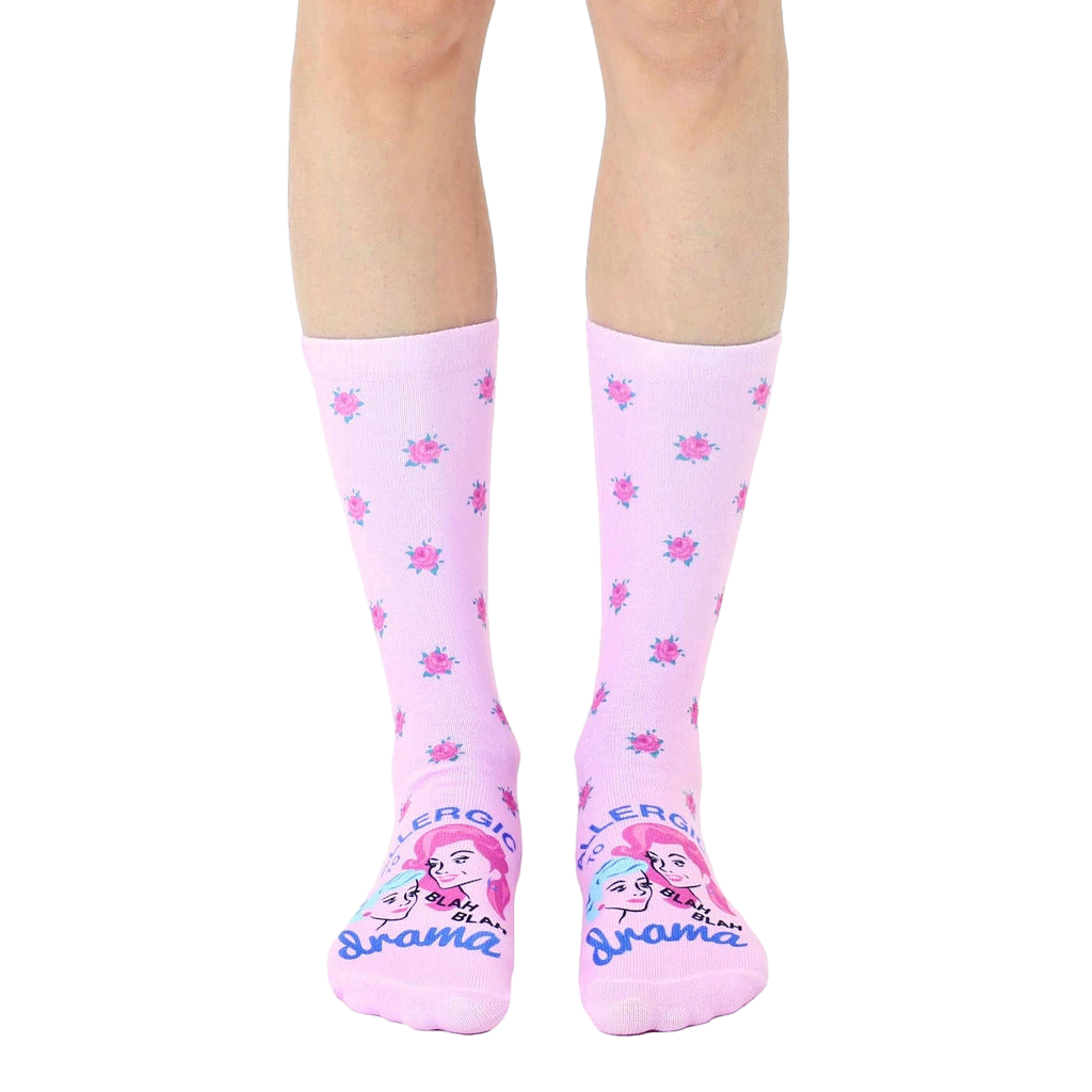 A pair of pastel pink crew socks with a scattered rose pattern and 'Allergic To Drama' graphic featuring two illustrated faces. [lf]