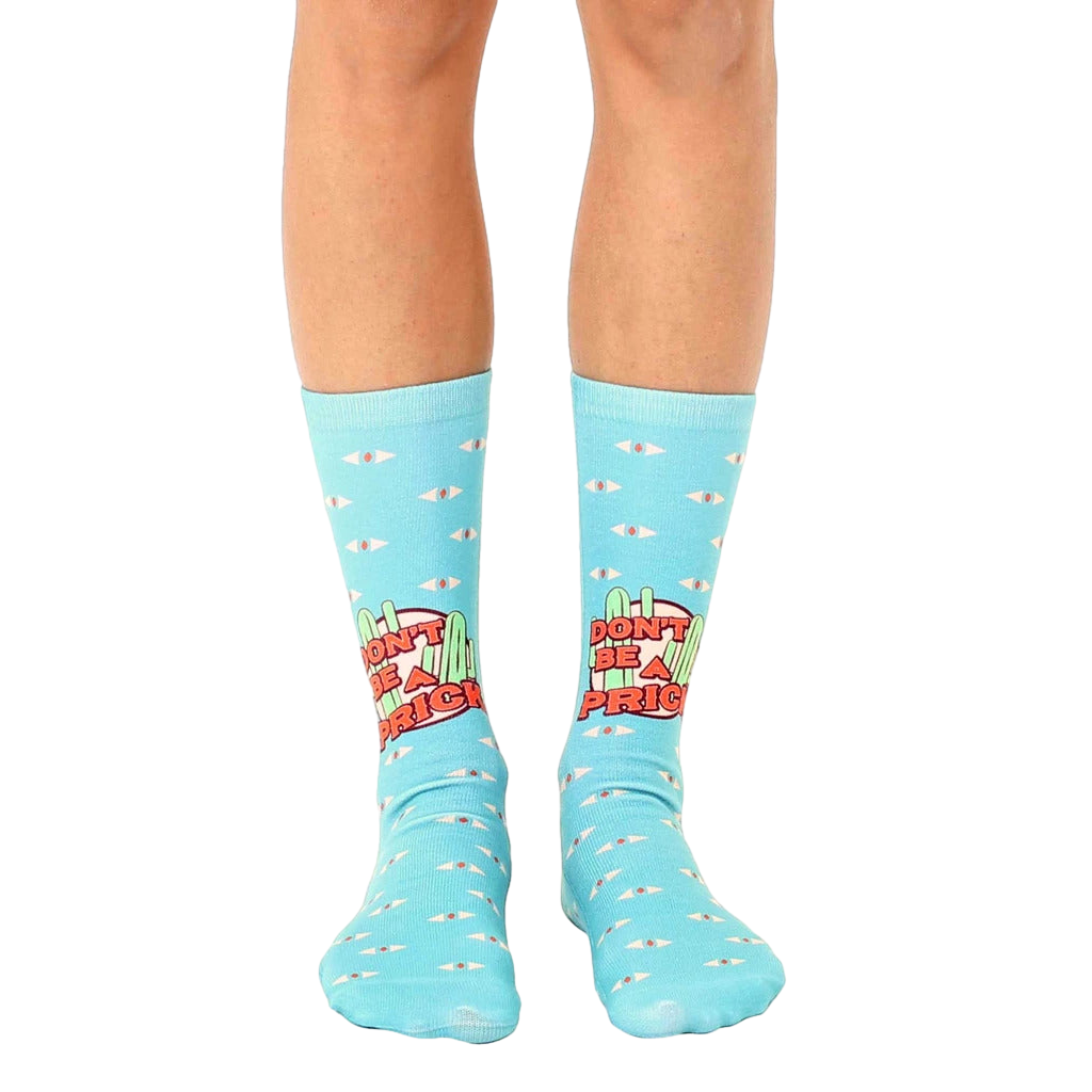 A pair of light blue crew socks with a 'Don't Be A Prick' slogan, cacti, and small eye illustrations. [lf]