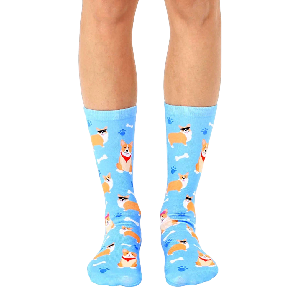 A pair of light blue crew socks featuring a repeating pattern of cute corgis wearing sunglasses, bandanas, and bows, alongside bones and paw prints. [lf]
