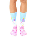 A pair of crew socks showing a birthday cake design.  The socks are light blue, pink, and purple with sprinkles and candles. [lf]