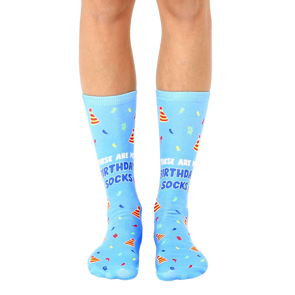 A pair of light blue crew socks with a birthday theme. They feature party hats, confetti, and the text 'These are my birthday socks.' [lf]