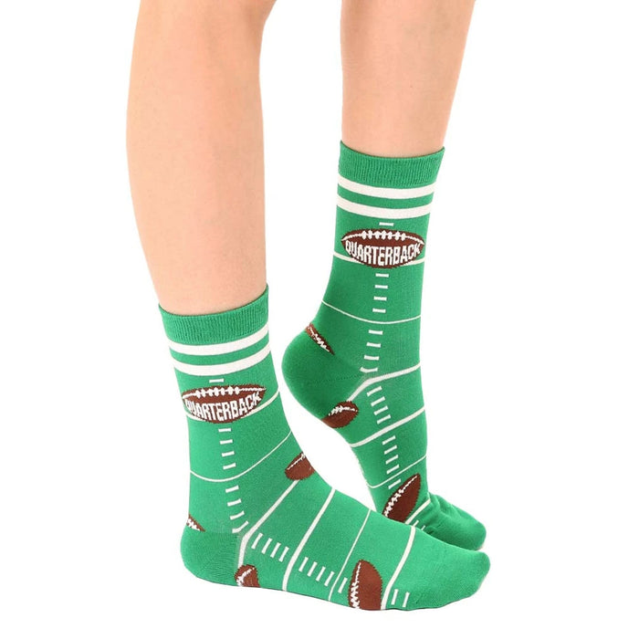 A pair of green baby socks with white stripes at the top and non-skid soles. The socks have a football field pattern with white yard markers and end zones. Brown footballs are centered on each sock. The word 