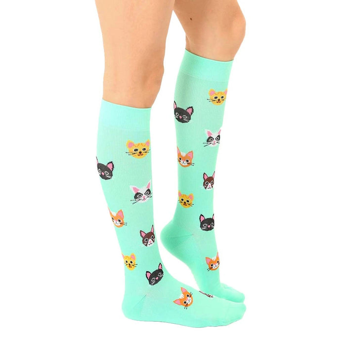 A pair of mint green knee-high socks with a pattern of cartoon cat faces in various colors.