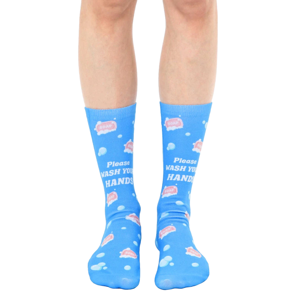 Pair of light blue crew socks featuring a playful design of pink soap bars and bubbles, with the text 'Please Wash Your Hands' repeated. [lf]