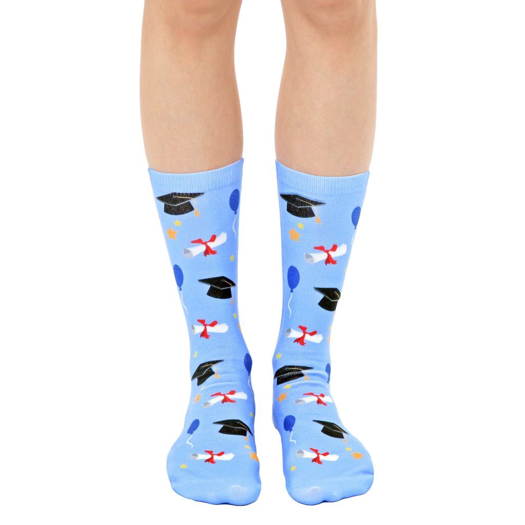 Pair of light blue crew socks with a graduation theme. The design includes black graduation caps, diplomas, and colorful balloons. [lf]