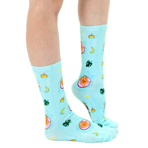A pair of blue socks with a pattern of cartoon sloths in inner tubes floating in a blue sea. There are also palm leaves, coconuts, and crescent moons in the pattern.