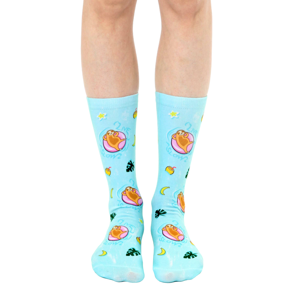A pair of light blue crew socks with a fun design. Sloths lounging on pink inner tubes in a pool are repeated against a tropical background with yellow stars, moons, and leaves. [lf]