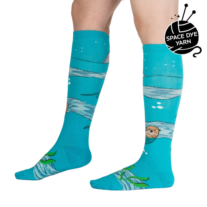 A pair of blue knee-high socks with an illustration of an otter in the water on each sock. The otters are swimming on their backs and holding a fish in their paws. There are also fish, a puffin, and snowflakes in the illustration.