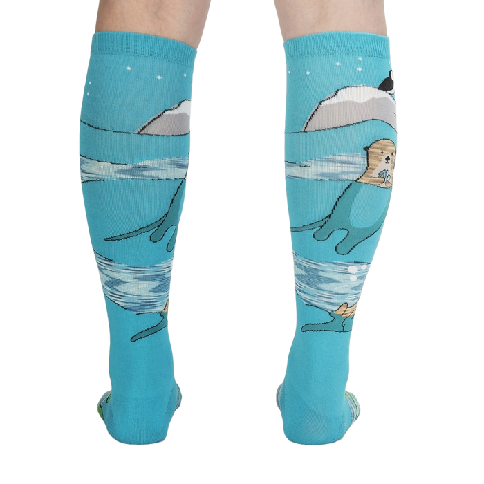 A pair of blue knee-high socks with an illustration of an otter in the water on each sock. The otters are swimming on their backs and holding a fish in their paws. There are also fish, a puffin, and snowflakes in the illustration.