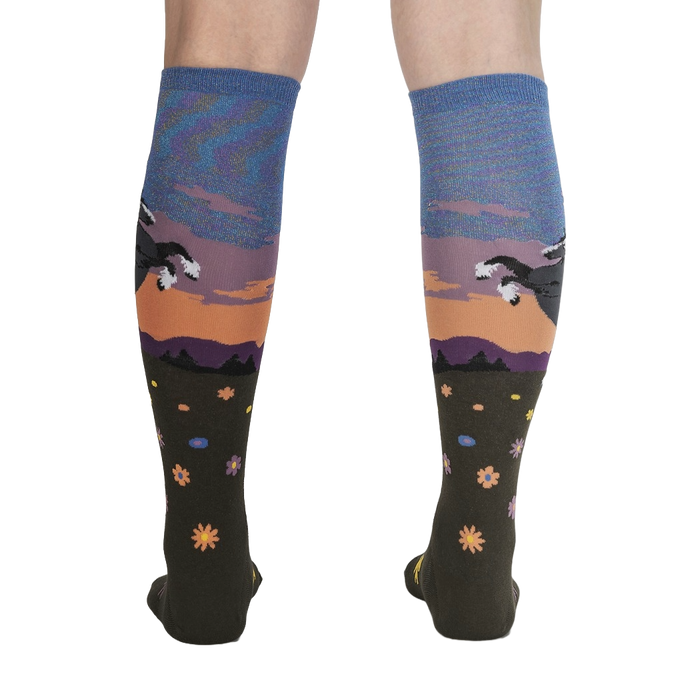 A pair of dark green knee-high socks with a colorful pattern of a mountain landscape with two rabbits running through a field of flowers at sunset.