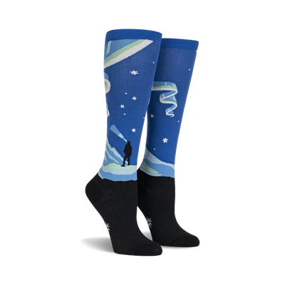 women's blue and black knee-high northern lights glowing socks illuminate the night with stars, mountains, and aurora borealis design.  