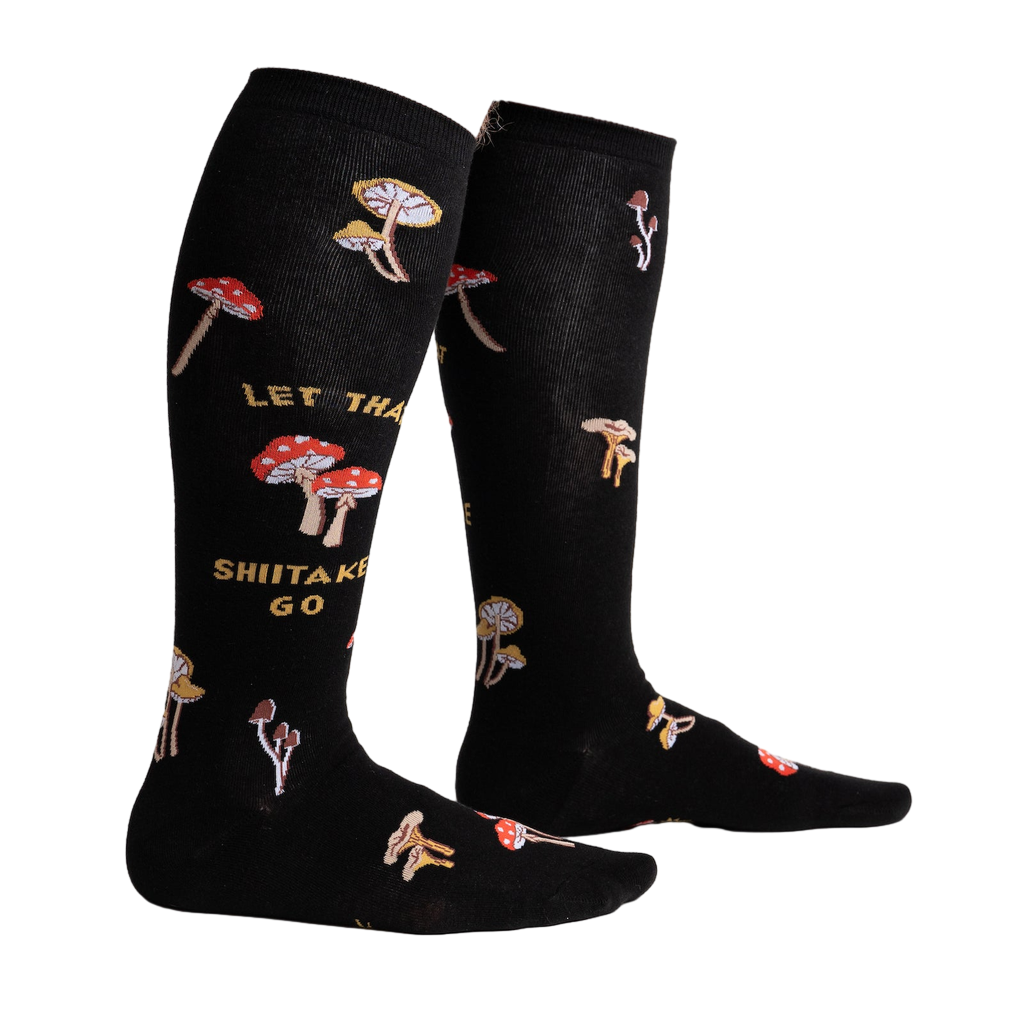 Lifestyle shot showing the 'Let That Shiitake Go' socks on a person's legs. The socks are black with a mushroom and text pattern.