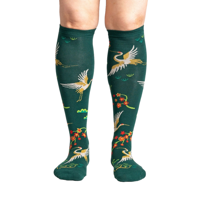 A pair of forest green knee-high socks with a repeating pattern of red-crowned cranes and cherry blossoms on a black background.