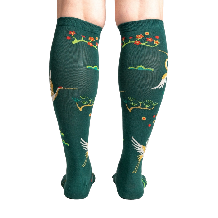 A pair of forest green knee-high socks with a repeating pattern of red-crowned cranes and cherry blossoms on a black background.