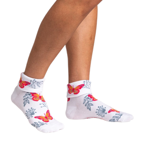 a woman is wearing white ankle socks with a butterfly print. the socks are on her feet.  the socks are a front view and the image is a side view. the butterflies are orange, red, and pink. the socks also have gray foliage.