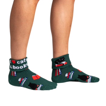 front view of a person wearing green ankle socks with a black cat, books and a red heart on them. one sock says, "i love cats and books."